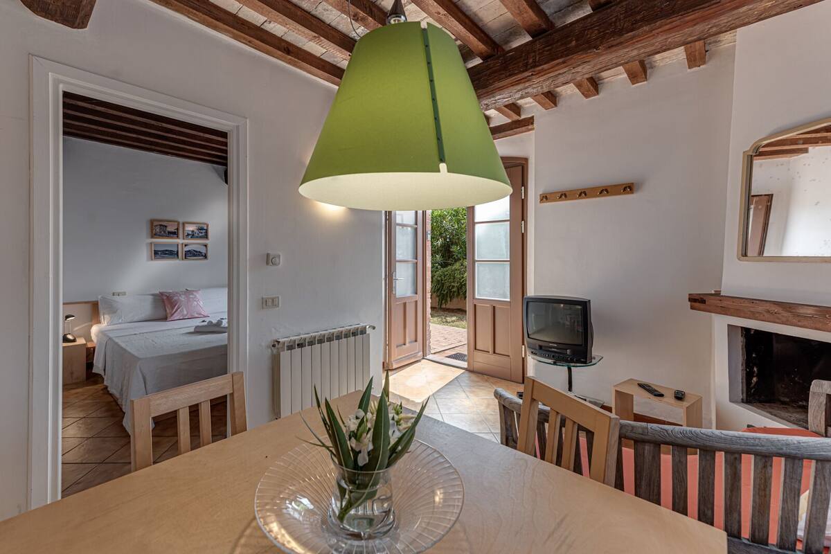 A cozy room with a wooden table, a green lamp, a bed with a white headboard, and a television on a glass stand.