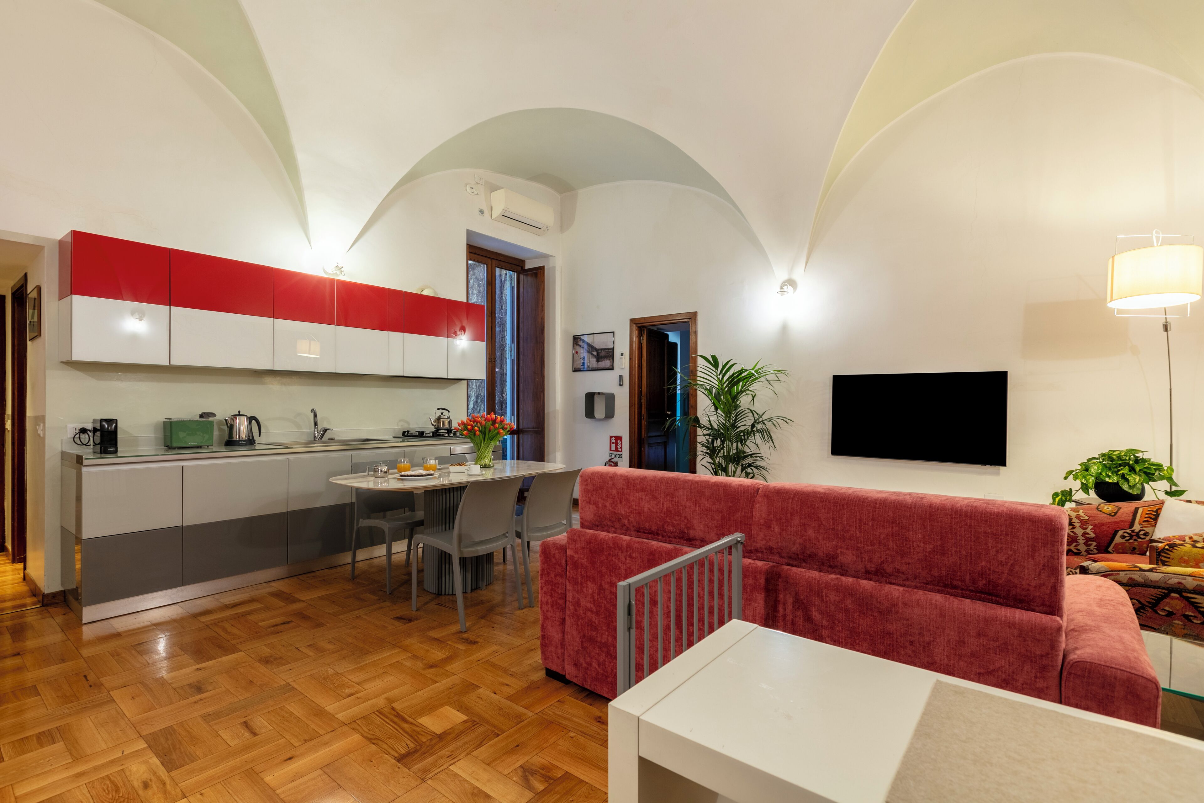 A modern kitchen with a dining area, a red sofa, and a TV mounted on the wall.