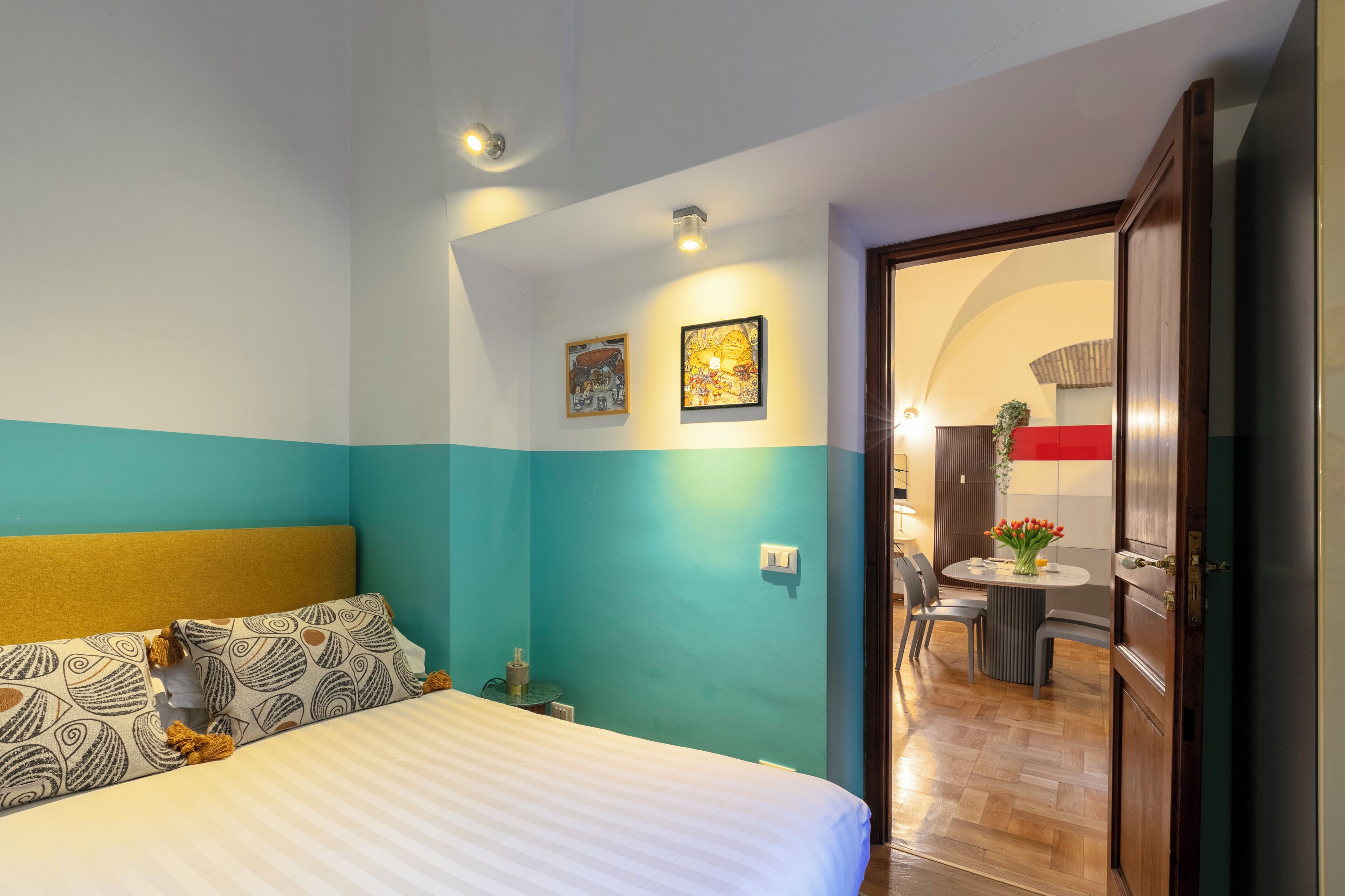A hotel room with a bed, a small table with a vase of flowers, and a door leading to a dining area.