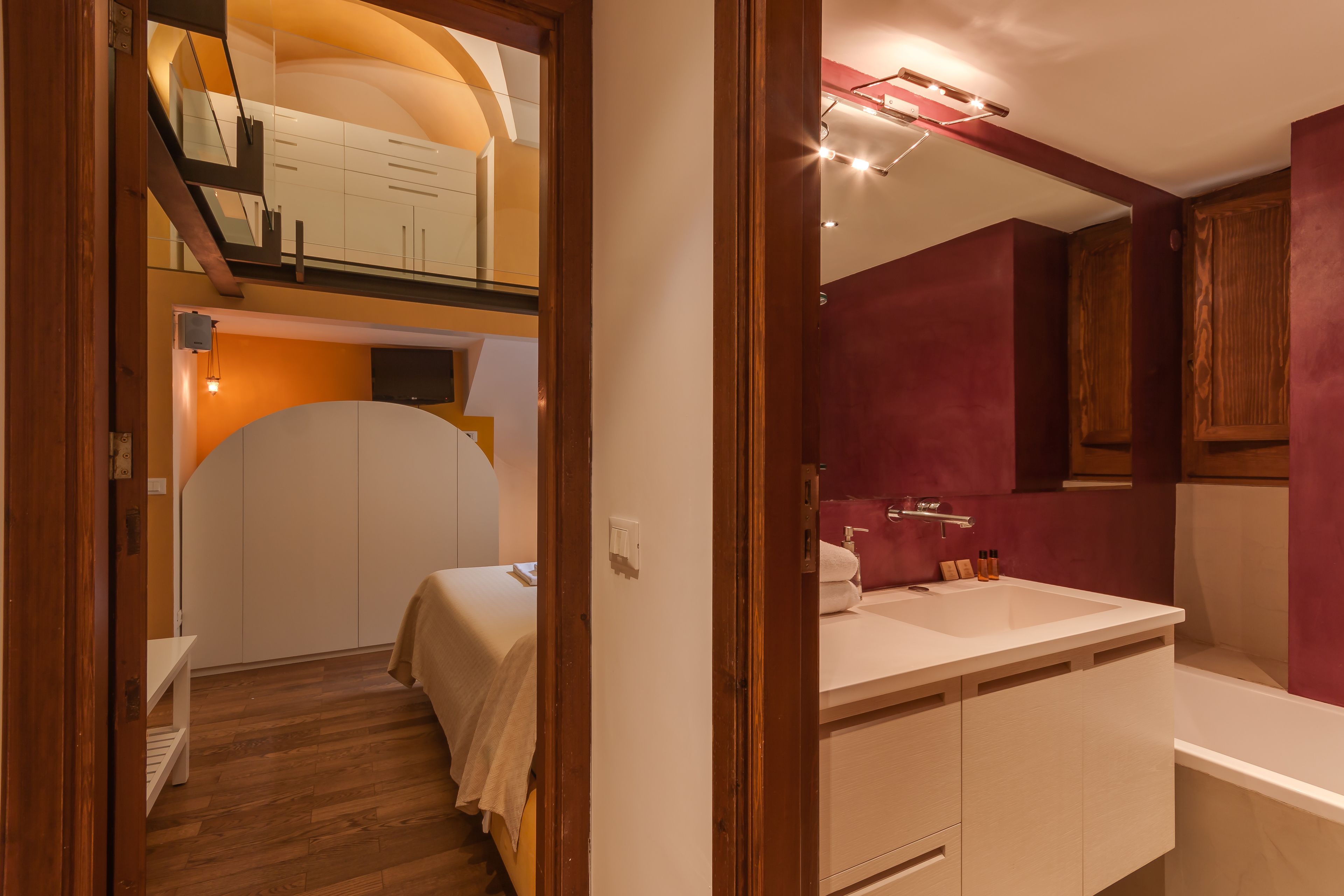 Ein Schlafzimmer mit Bett, Nachttisch und kleiner Ablage. Ein Fenster mit Vorhängen, eine Holztür und ein kleines Regal mit Büchern.
