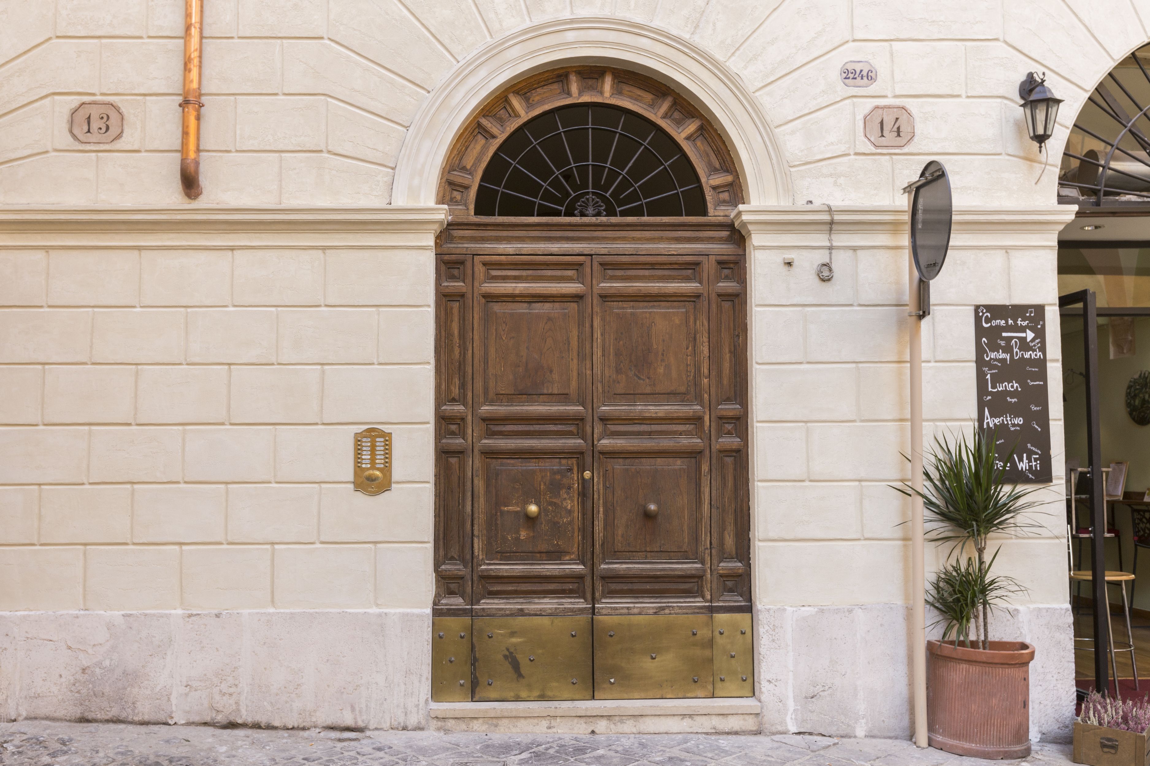 A large wooden double door with brass accents, flanked by two numbers on the wall, and a potted plant to the right.