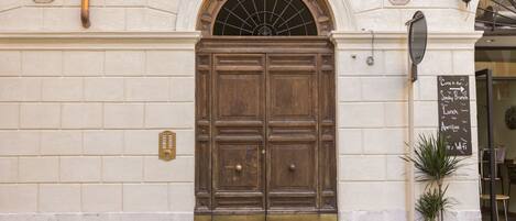 A large wooden double door with brass accents, flanked by two numbers on the wall, and a potted plant to the right.