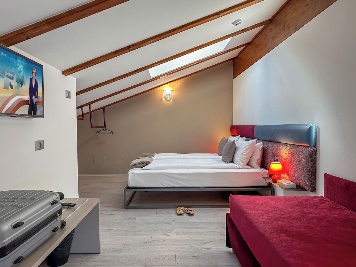 Quarto moderno com teto estilo loft, cama com roupa de cama branca, parede de destaque vermelha e uma pequena mesa de cabeceira com abajur.