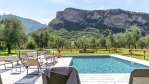 A swimming pool with lounge chairs and a wooden deck, surrounded by greenery and a mountain backdrop. - O-live Agriresort (Arco)