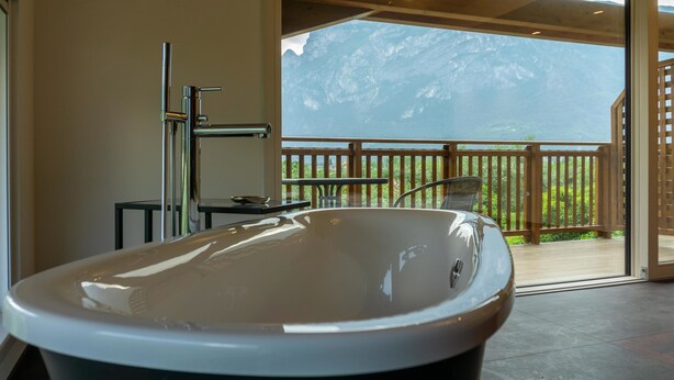A modern bathroom with a freestanding bathtub, a shower, and a view of a wooden deck and mountains. - O-live Agriresort (Arco)