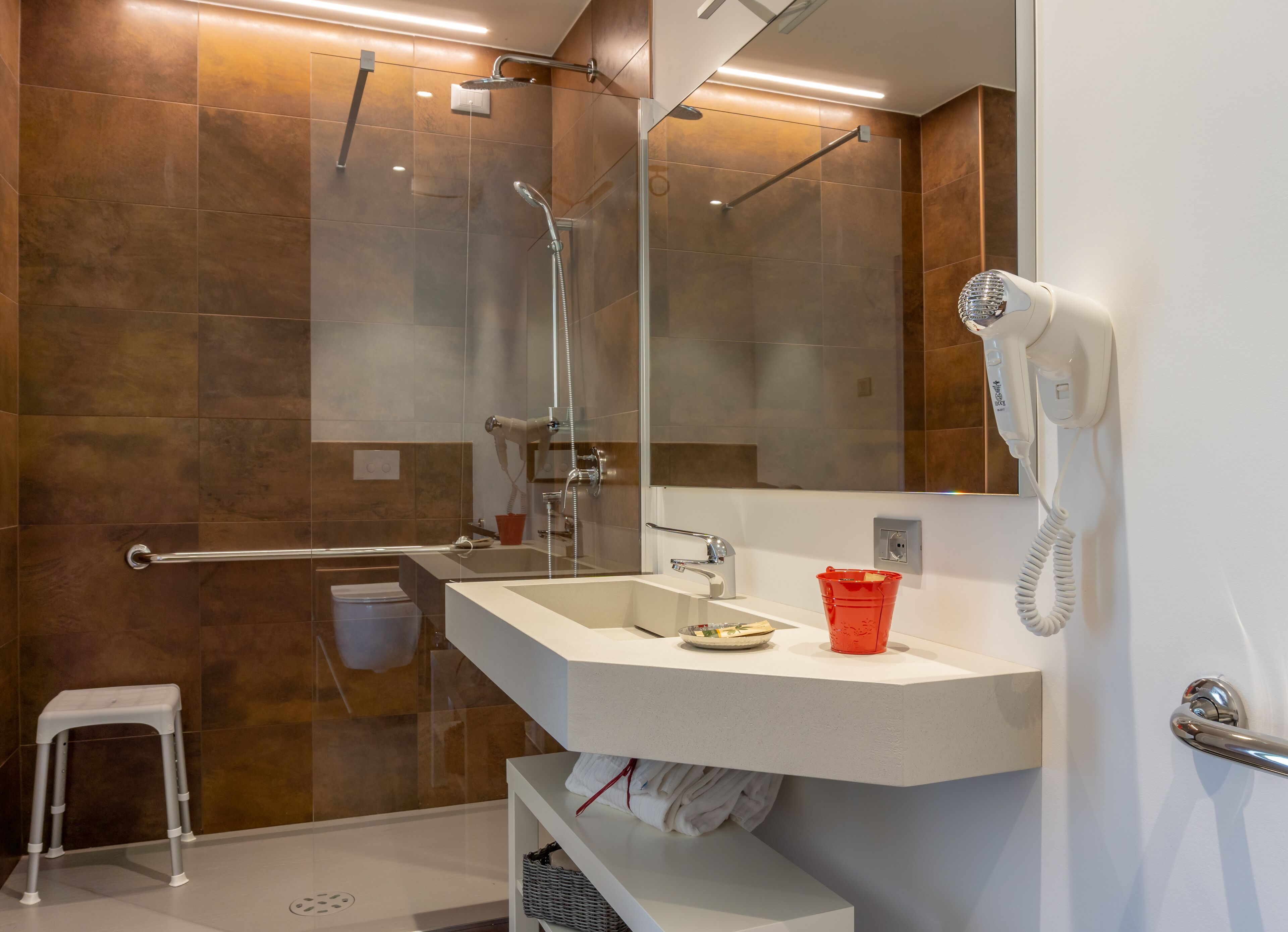A modern bathroom with a glass shower enclosure, a white sink, a red bucket, and a hairdryer mounted on the wall.