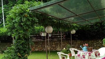 Un patio couvert avec des chaises et une table en plastique blanc, entouré de verdure et d’un mur de pierres.
