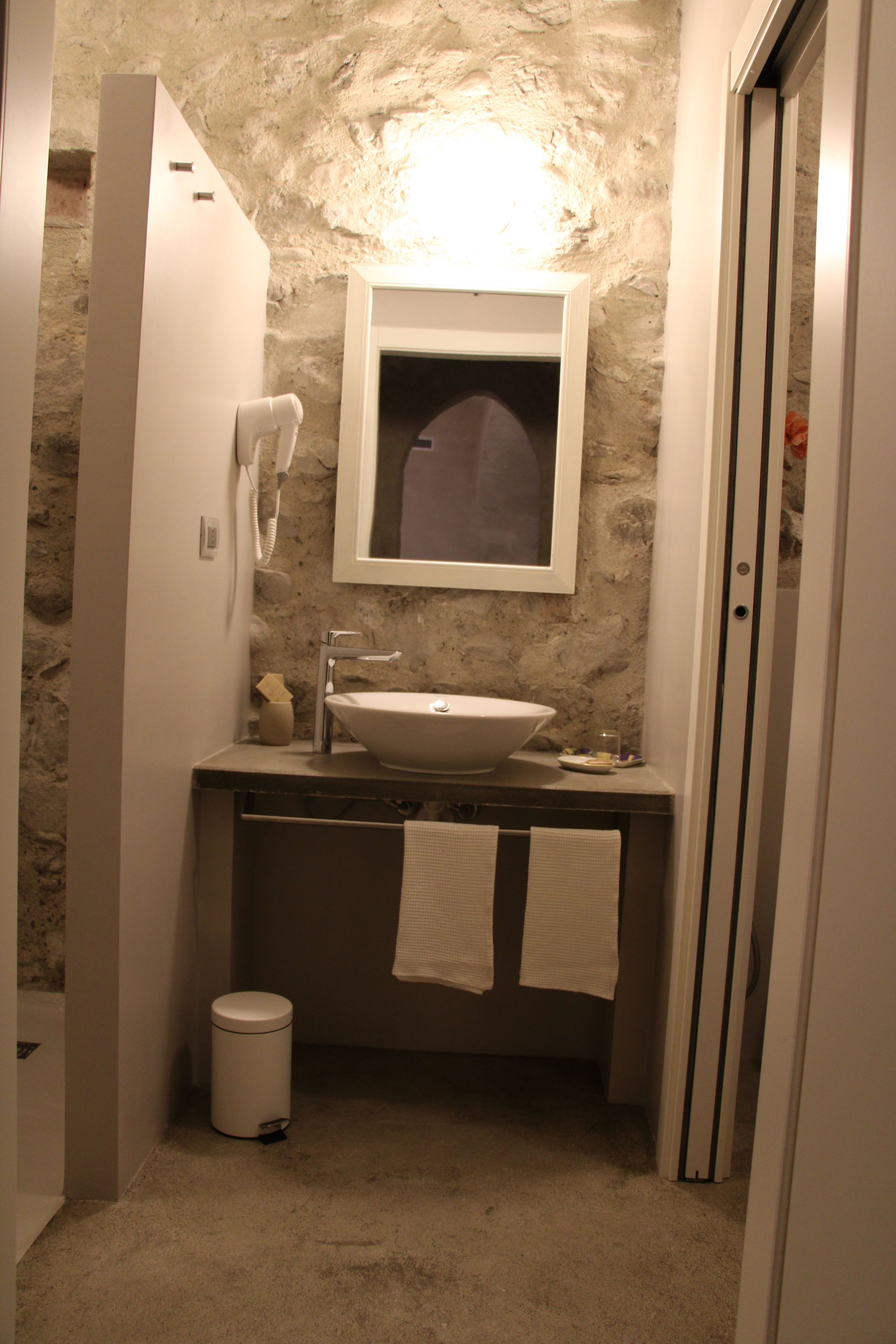 Un baño con pared de piedra, un lavabo blanco, un espejo, un perchero para toallas y un cubo de basura.