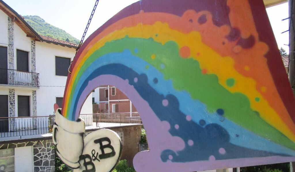 Une fresque colorée représentant un arc-en-ciel et une lettre « B » stylisée, peinte sur la façade d’un immeuble.