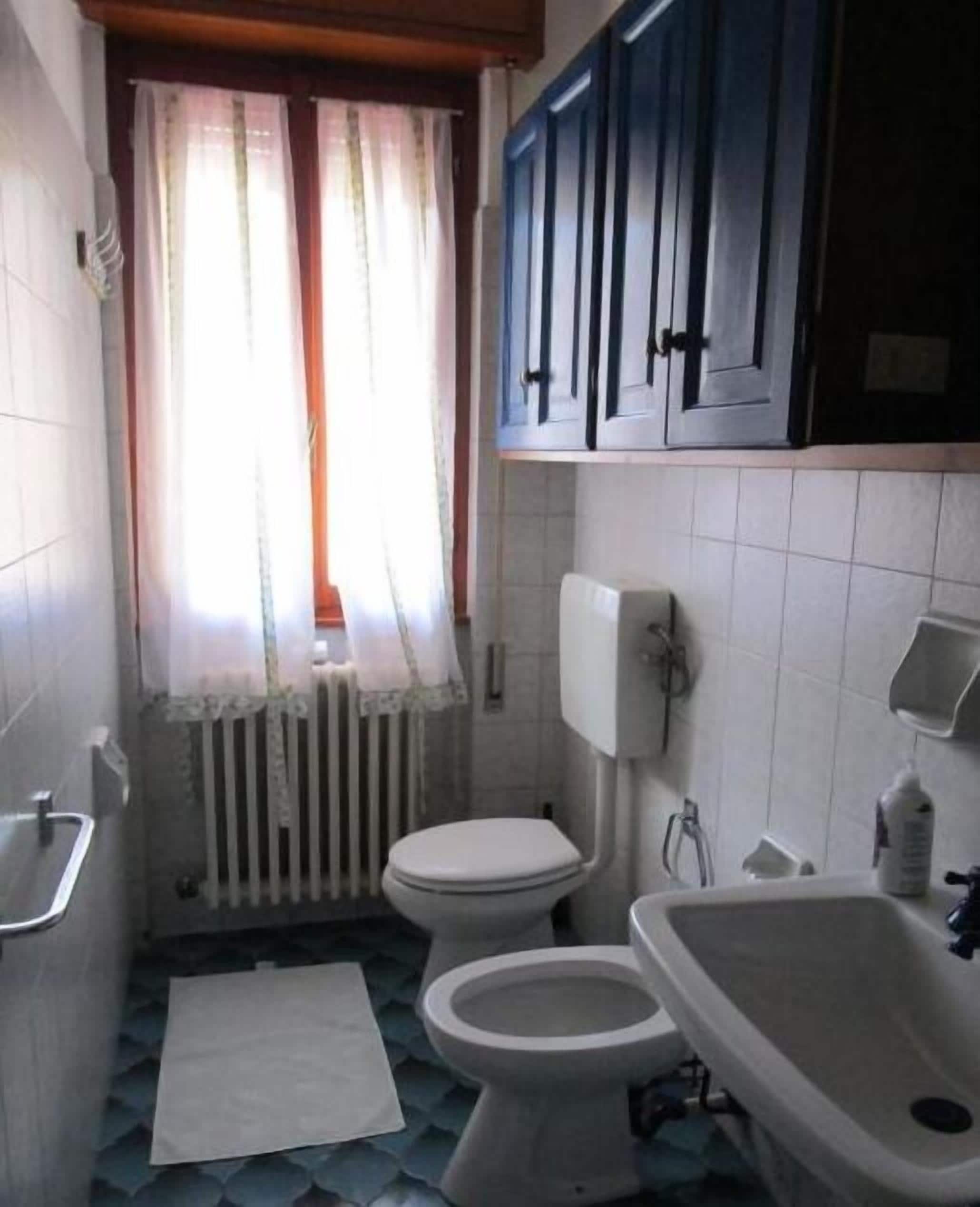 Ein kleines Badezimmer mit zwei Toiletten, einem Waschbecken, einem Heizkörper und einem Fenster mit Vorhängen.