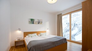 A bedroom with a wooden bed, a nightstand, a window with a view of snow-covered mountains, and wall art. - Appartements Berg 170 (Damüls)