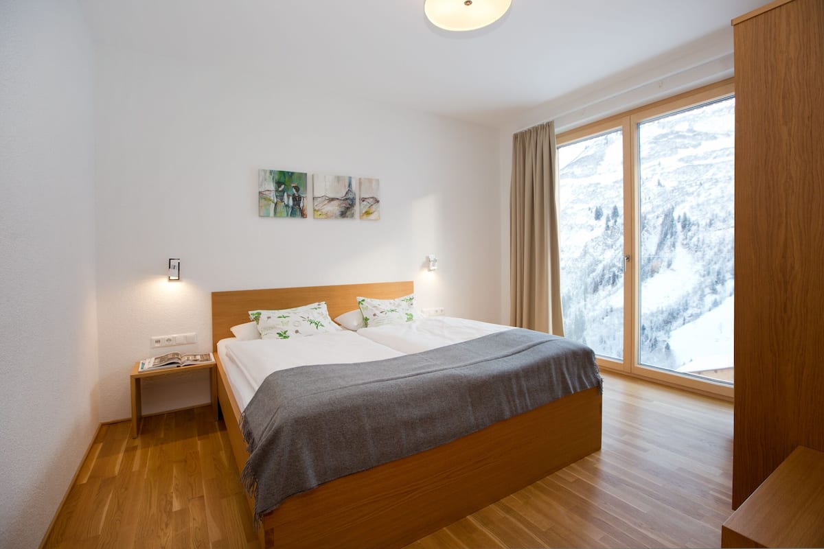 A bedroom with a wooden bed, a nightstand, a window with a view of snow-covered mountains, and wall art.