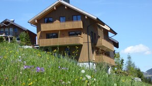 A modern two-story wooden house with balconies, surrounded by a lush green field with wildflowers. - Appartements Berg 170 (Damüls)