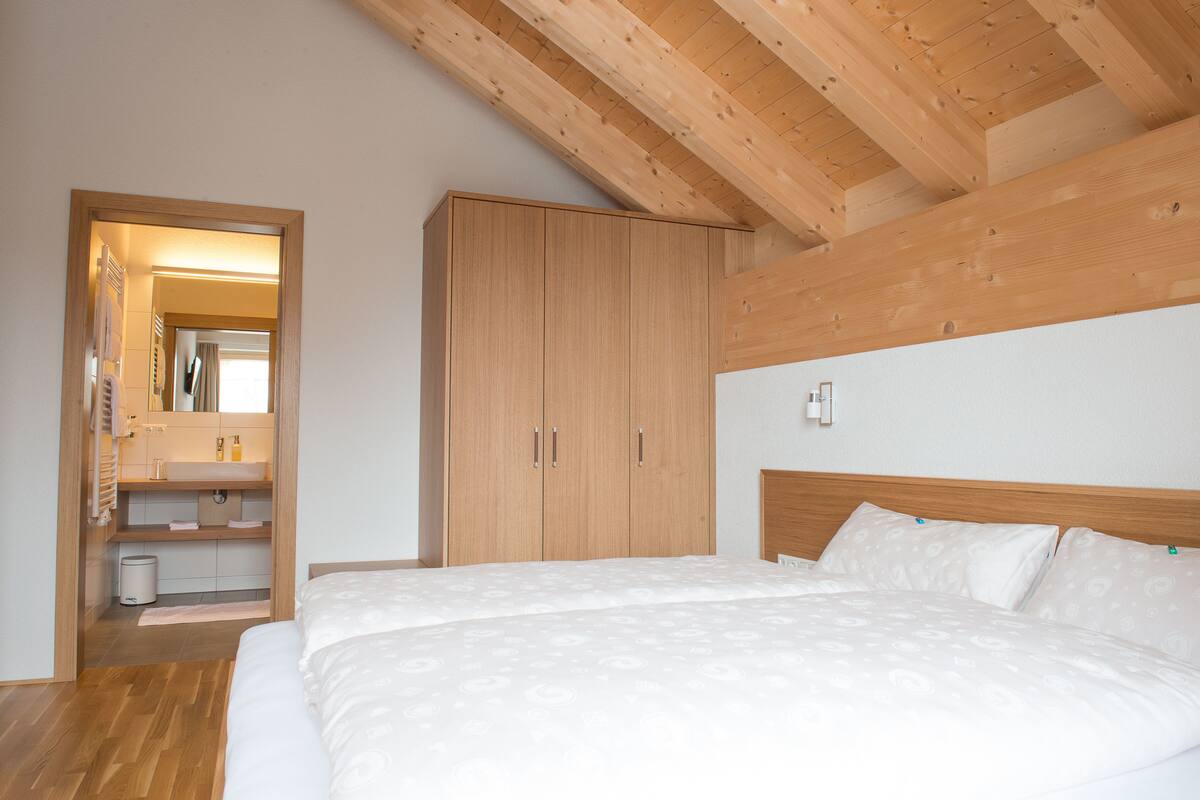 A bedroom with a wooden ceiling, a bed with white bedding, a wooden wardrobe, and a bathroom with a mirror and sink.