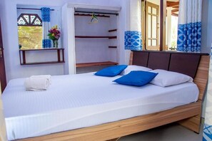 A single bed with a wooden frame, white bedding, and blue pillows. There is a towel on the bed, and a window with blue curtains and a hanging plant. - DiNi Galle (Galle)