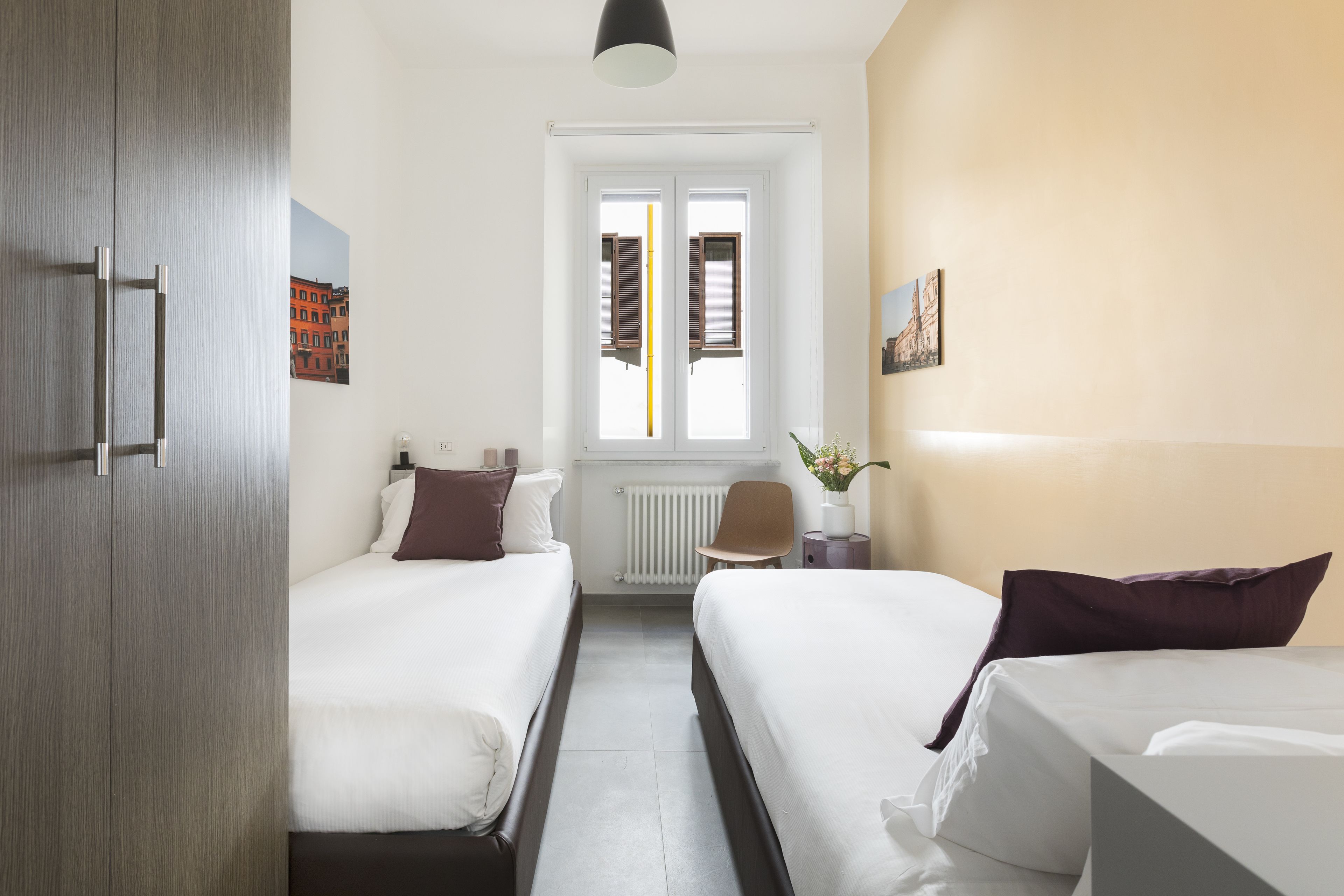 Een hotelkamer met twee bedden, een radiator, een raam met luiken, een stoel en een klein tafeltje met een vaas.