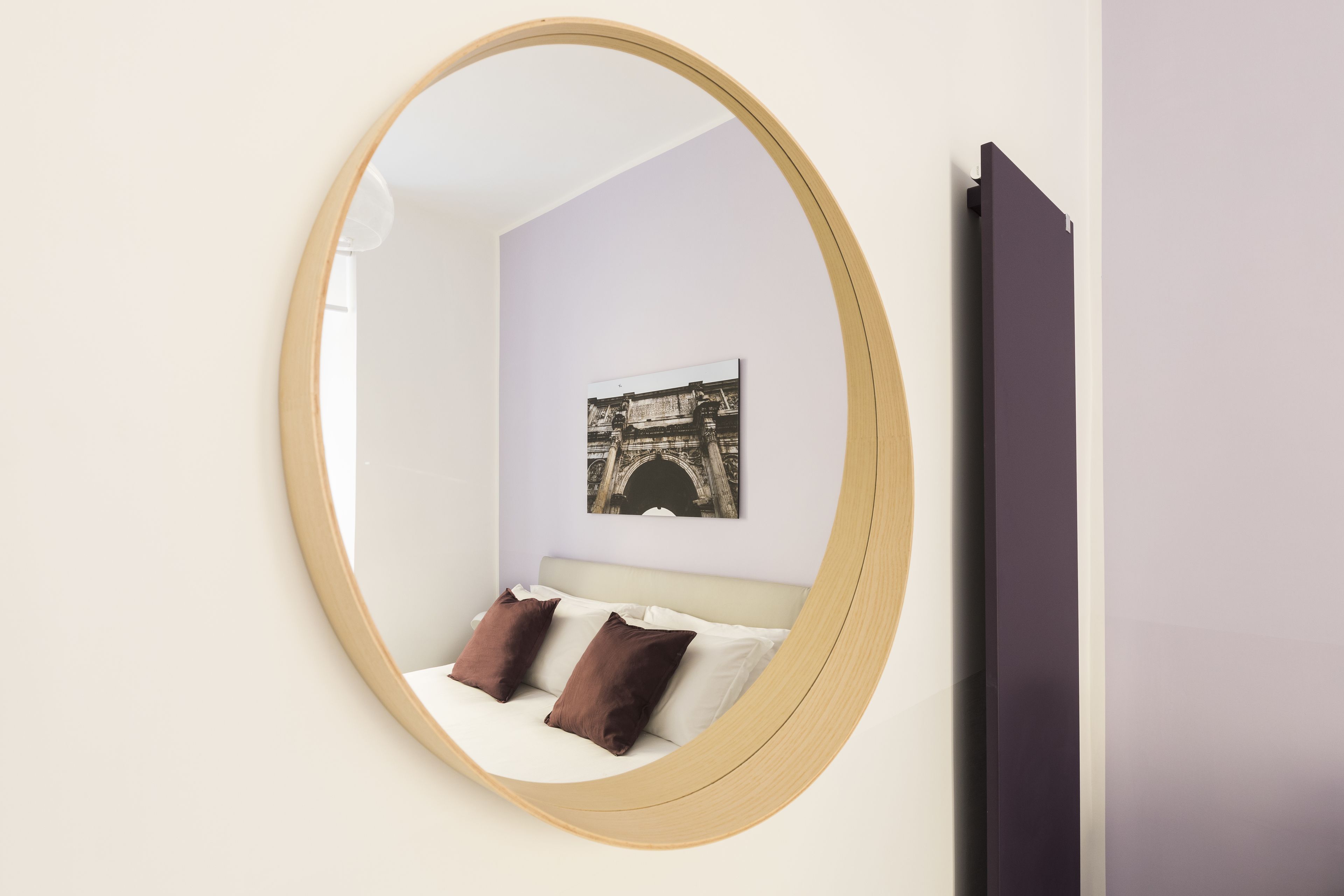 A bedroom with a round mirror, a bed with two pillows, a wall-mounted picture, and a dark-colored door.