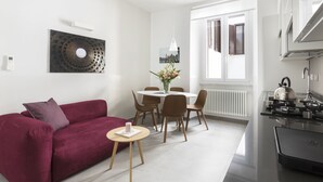 En moderne stue med en rød sofa, et rundt spisebord med stole, et lille sofabord med en bog og et køkkenområde med komfur og skabe.