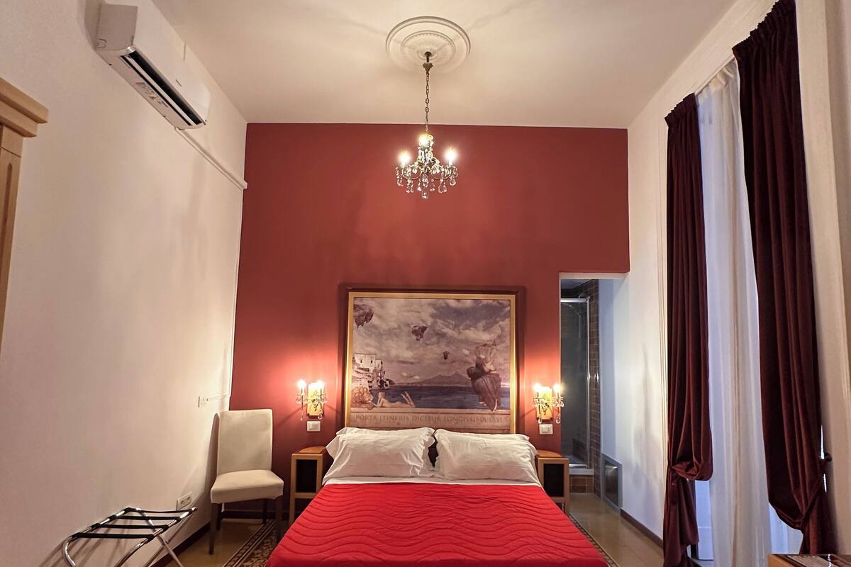 A hotel room with a large bed, a chandelier, a painting on the wall, and a window with curtains.