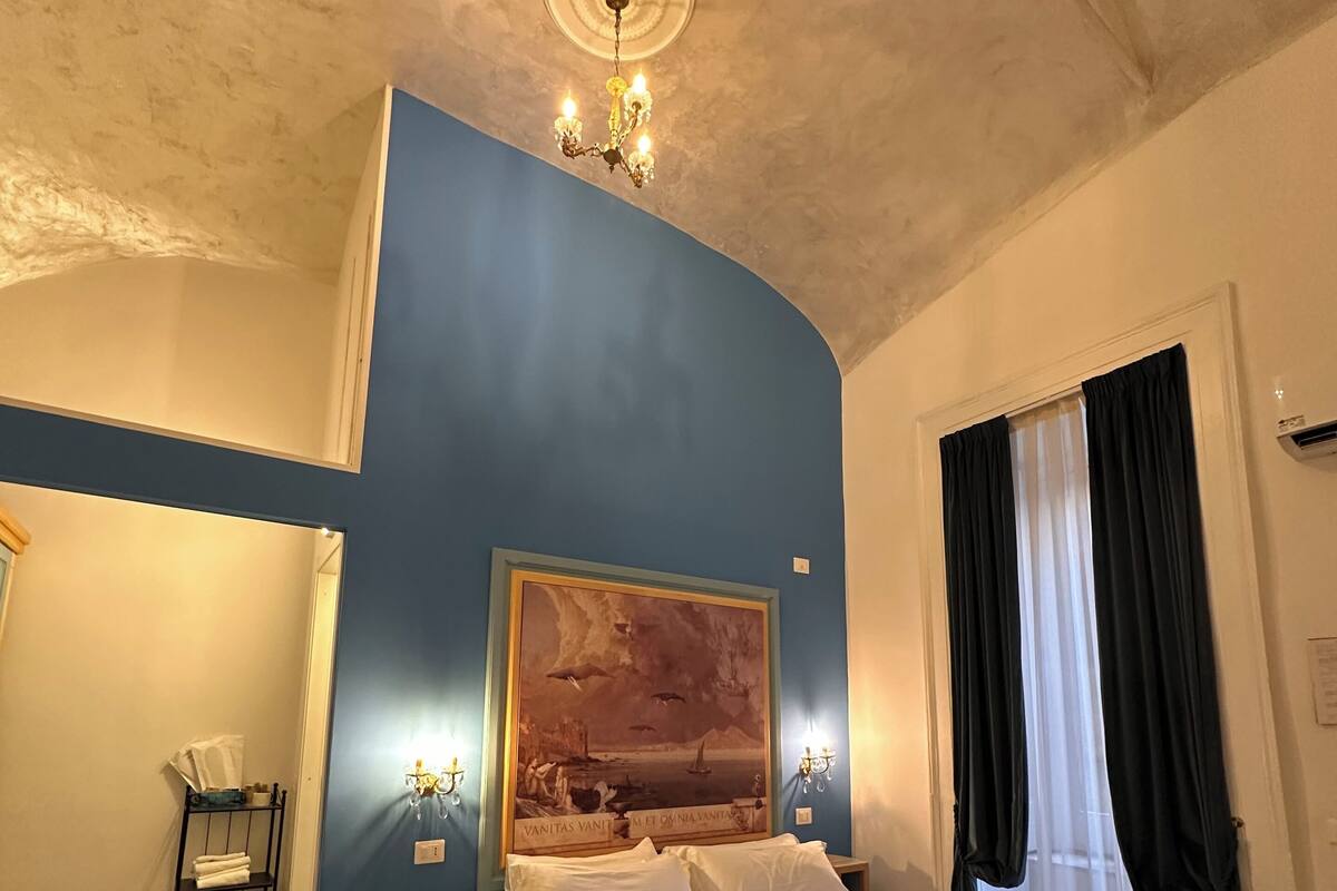 A bedroom with a bed, a painting on the wall, a chandelier, and a window with curtains.