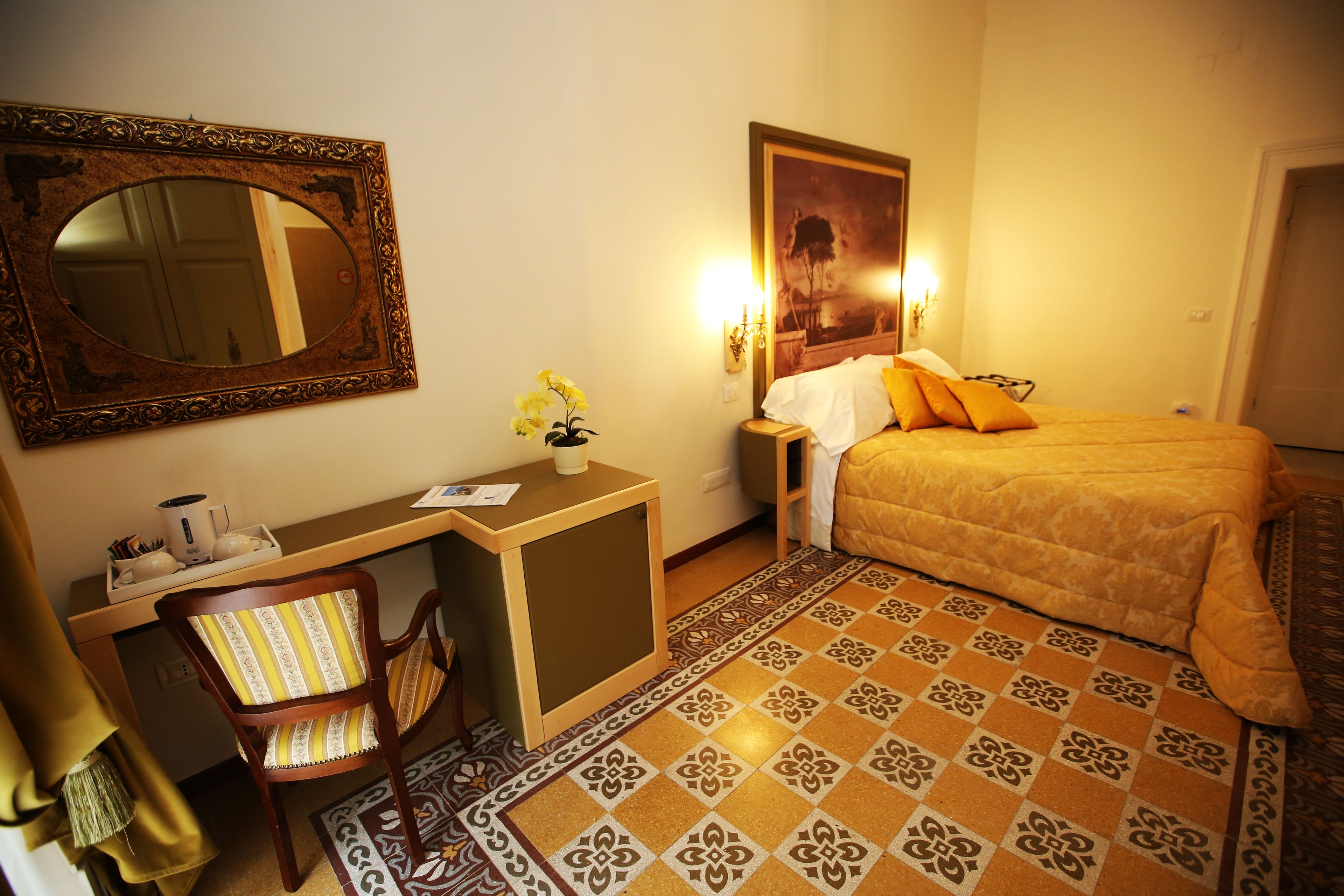 A hotel room with a bed, a desk with a chair, a mirror, a nightstand with a lamp, and patterned floor tiles.