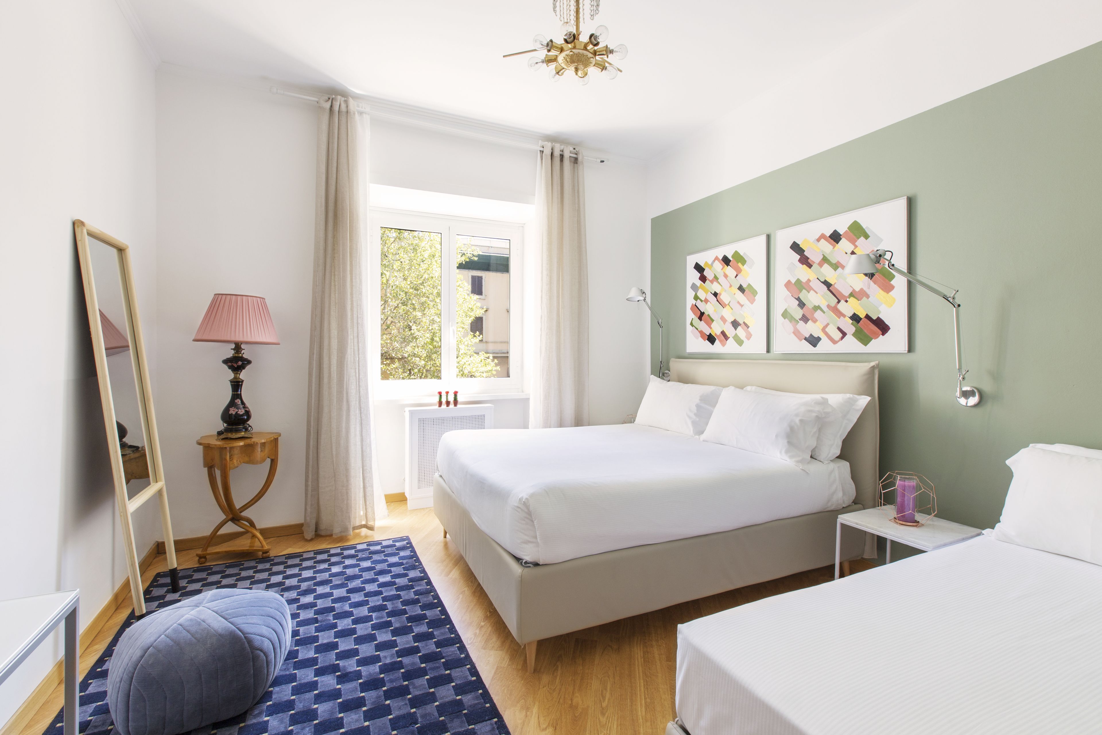 A hotel room with a large bed, a smaller bed, a bedside table with a lamp, a mirror, a window with curtains, and artwork on the wall.