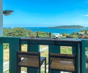 A balcony with two wooden chairs, a table with a candle, a small Eiffel Tower model, and a view of the ocean and distant hills.