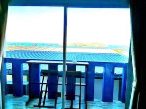 A room with a view of the ocean through large windows, a wooden deck, and a blue railing.