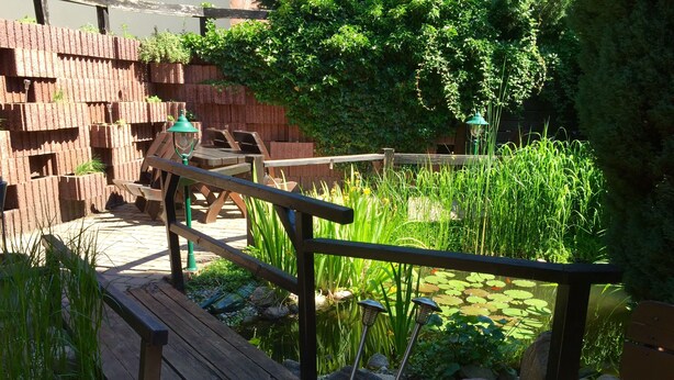 A wooden deck with a metal railing, a pond with lily pads, and a brick wall with greenery. - Pension Alabama (Lutherstadt Wittenberg)