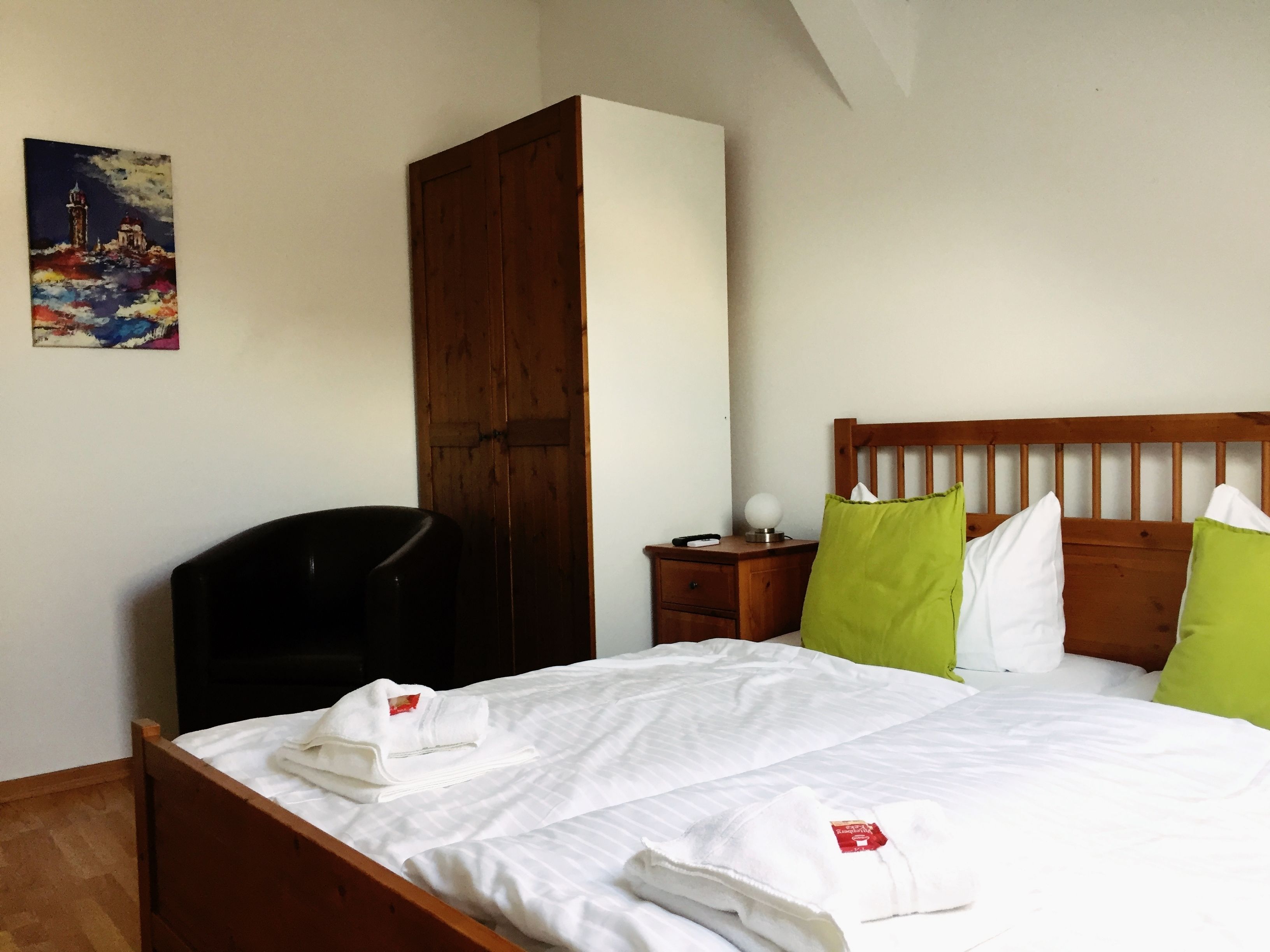 Una cama bien hecha con sábanas blancas, cabecera de madera y almohadas verdes y blancas. Se ve un armario de madera y una mesita de noche con una lámpara y un control remoto.