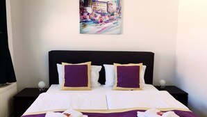 A neatly made bed with white linens and purple accents, two white towels folded on the bed, and a painting of a cityscape on the wall. - Pension Alabama (Lutherstadt Wittenberg)