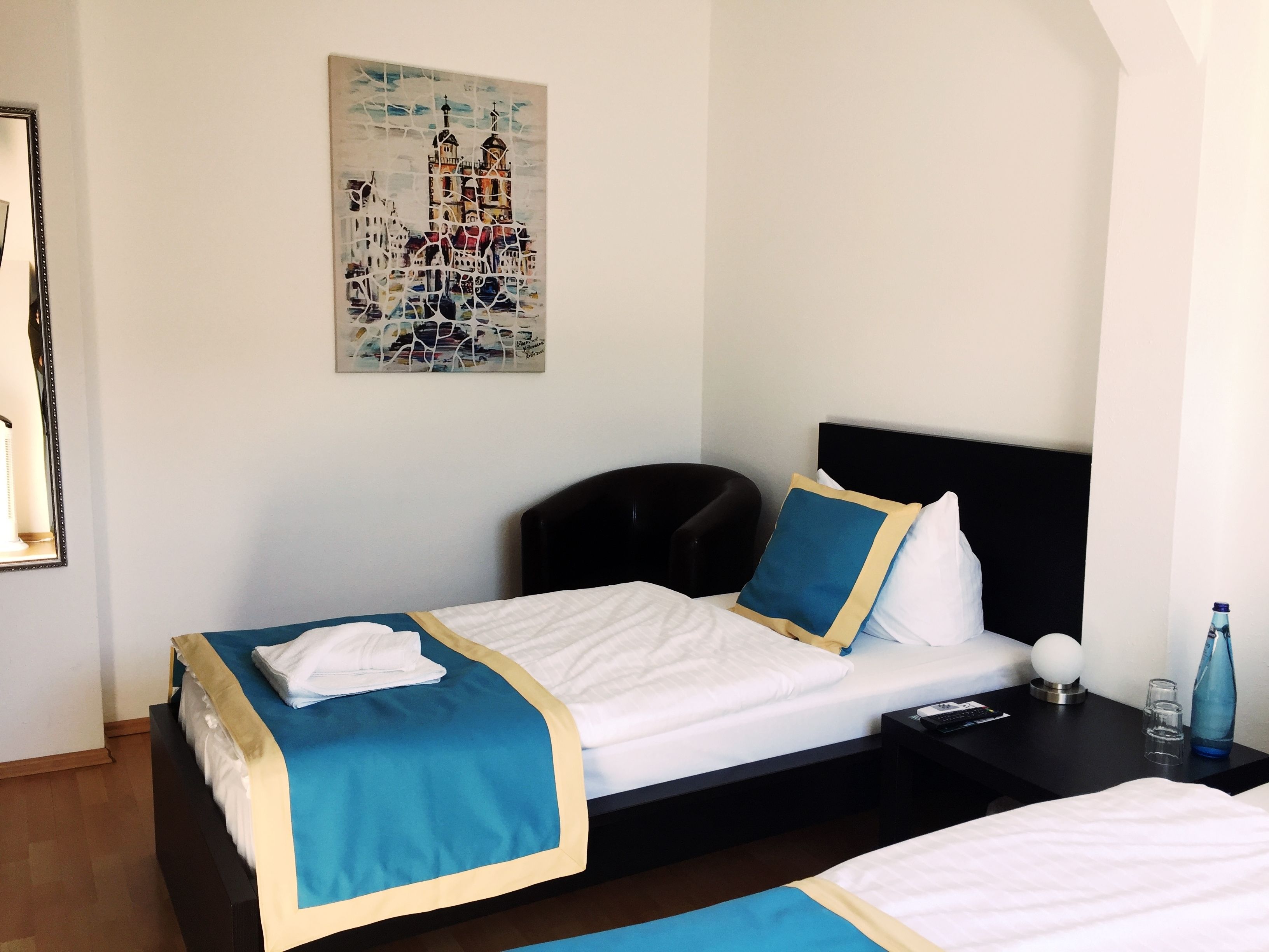 Una habitación de hotel con una cama, una mesita de noche, una lámpara, una botella de agua y un cuadro en la pared.