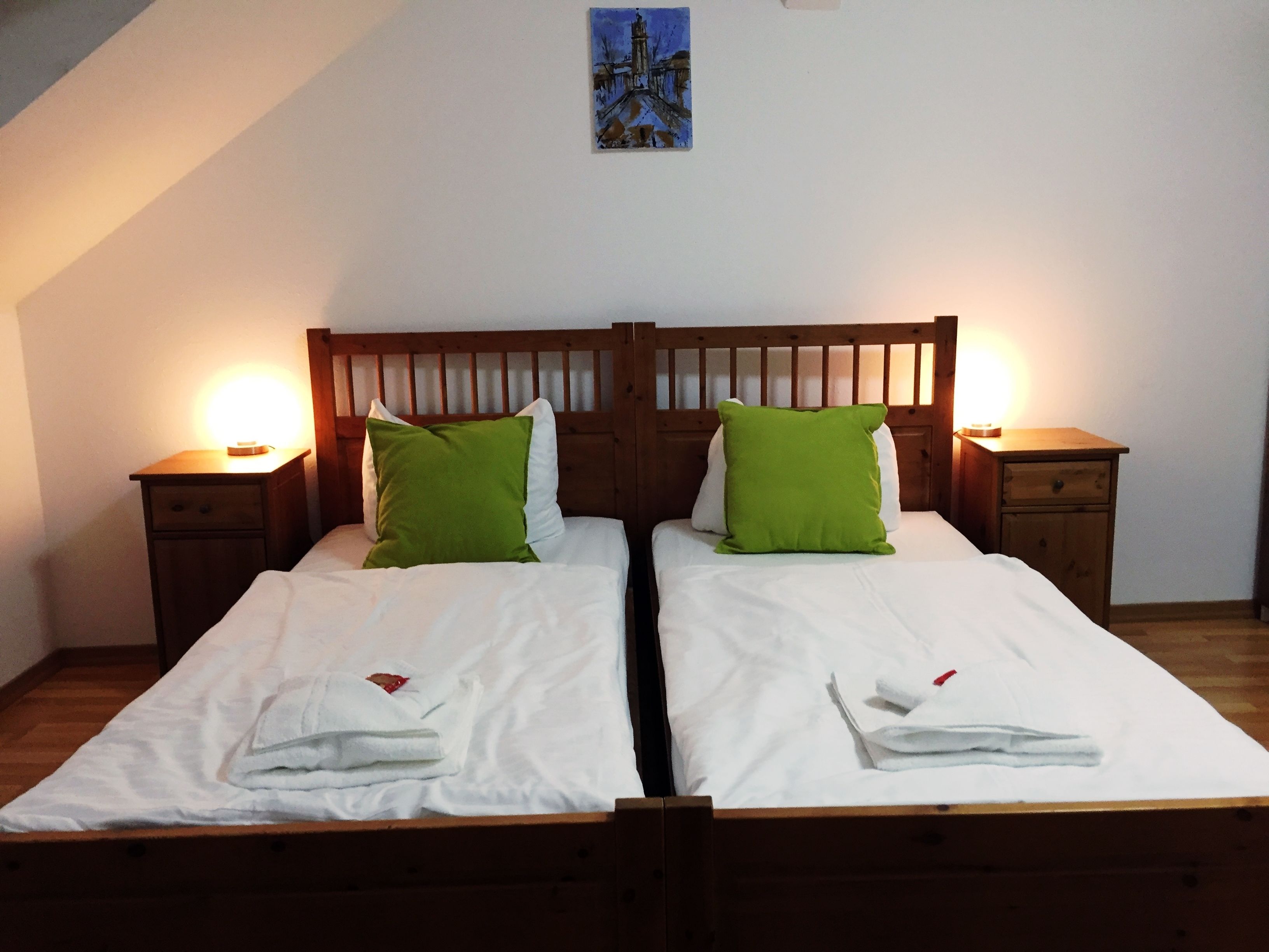 A double bed with white linens and green pillows, flanked by wooden nightstands with lamps, in a room with a sloped ceiling and a painting above the bed.