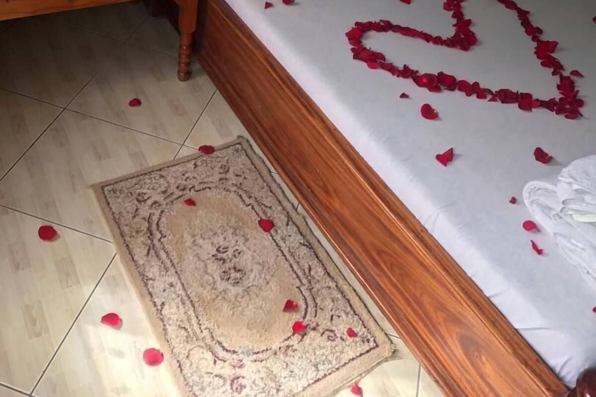A neatly made bed with rose petals arranged in a heart shape, a wooden nightstand with fruit and water bottles, and a patterned rug on the floor.