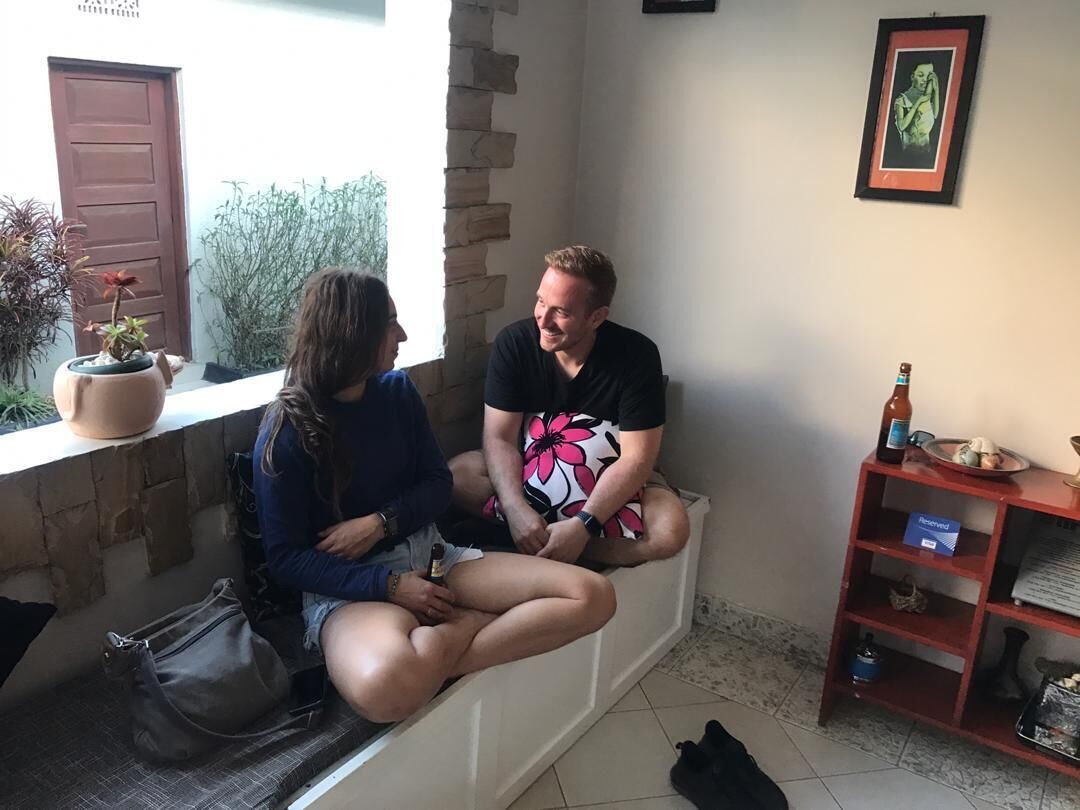 Two people sitting on a bench in a room with a stone wall, a window, and a shelf with items.