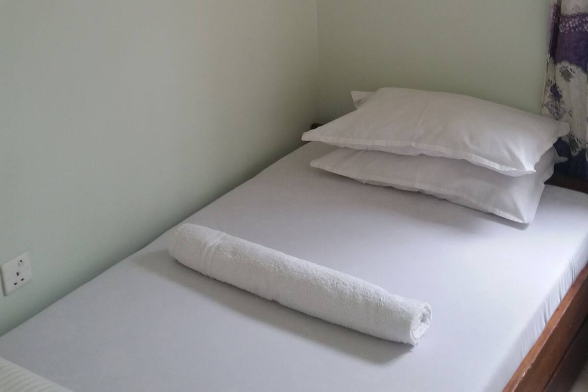 A single bed with white bedding and a rolled white towel, a white hanging canopy, and a purple curtain partially visible.