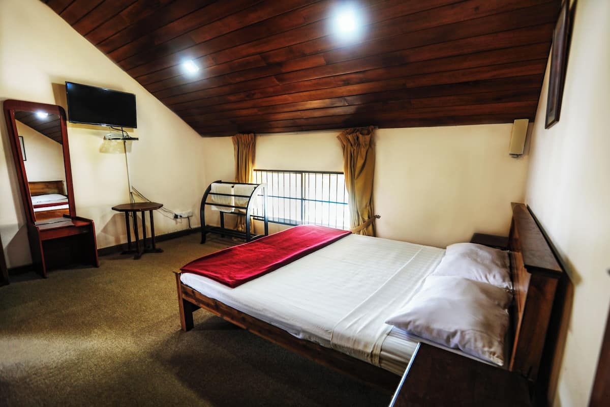 Ein Schlafzimmer mit Holzbalkendecke, einem Bett mit roter Decke, einem Schreibtisch mit Stuhl, einem an der Wand befestigten Fernseher und einem Fenster mit Vorhängen.
