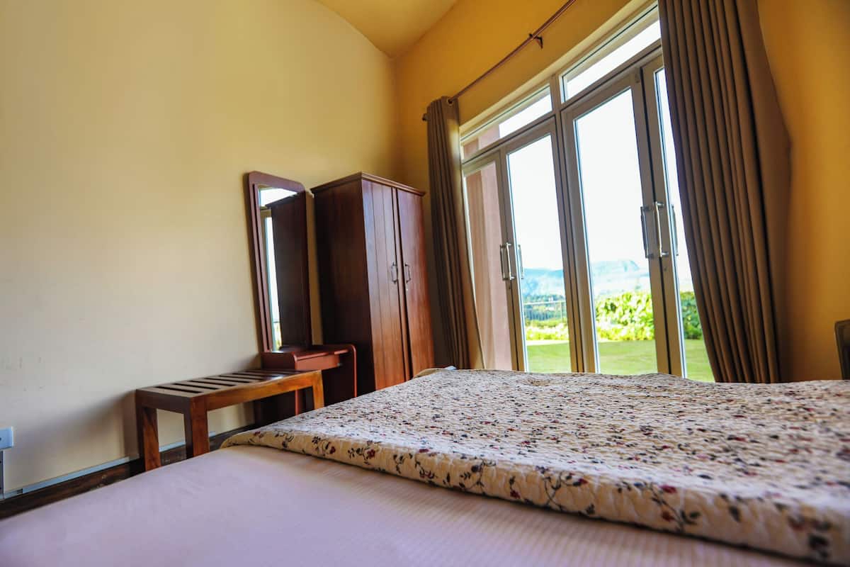 Ein Schlafzimmer mit einem großen Bett, einem Holztisch, einem Kleiderschrank und einer Glastür, die auf einen Balkon mit Blick ins Grüne führt.