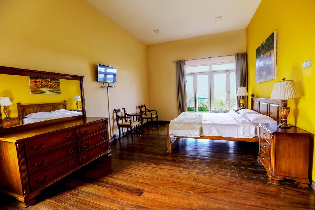 Ein Hotelzimmer mit einem großen Bett, einer Holzkommode, einem Schreibtisch mit Stühlen, einem Fernseher und einem Fenster mit Vorhängen.