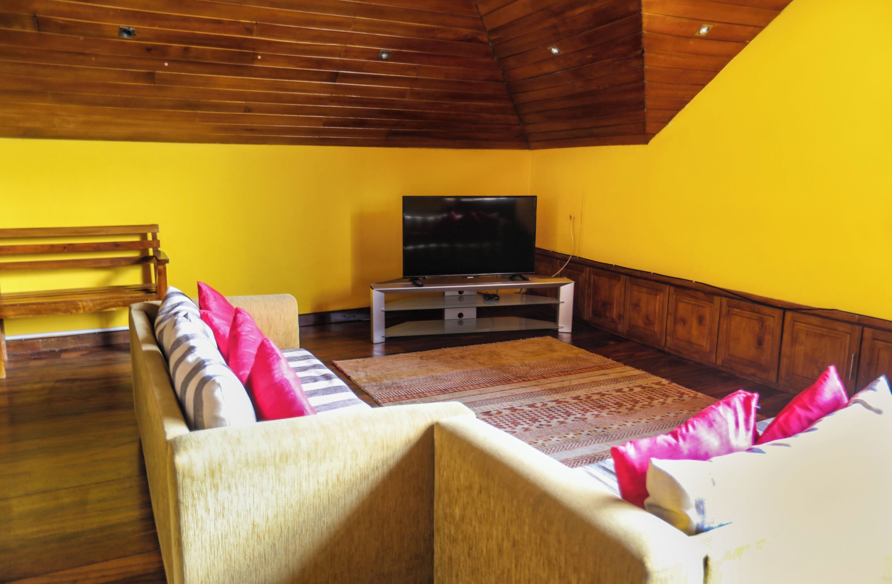 A living room with a wooden ceiling, yellow walls, a sofa with pillows, a television on a stand, and a patterned rug.