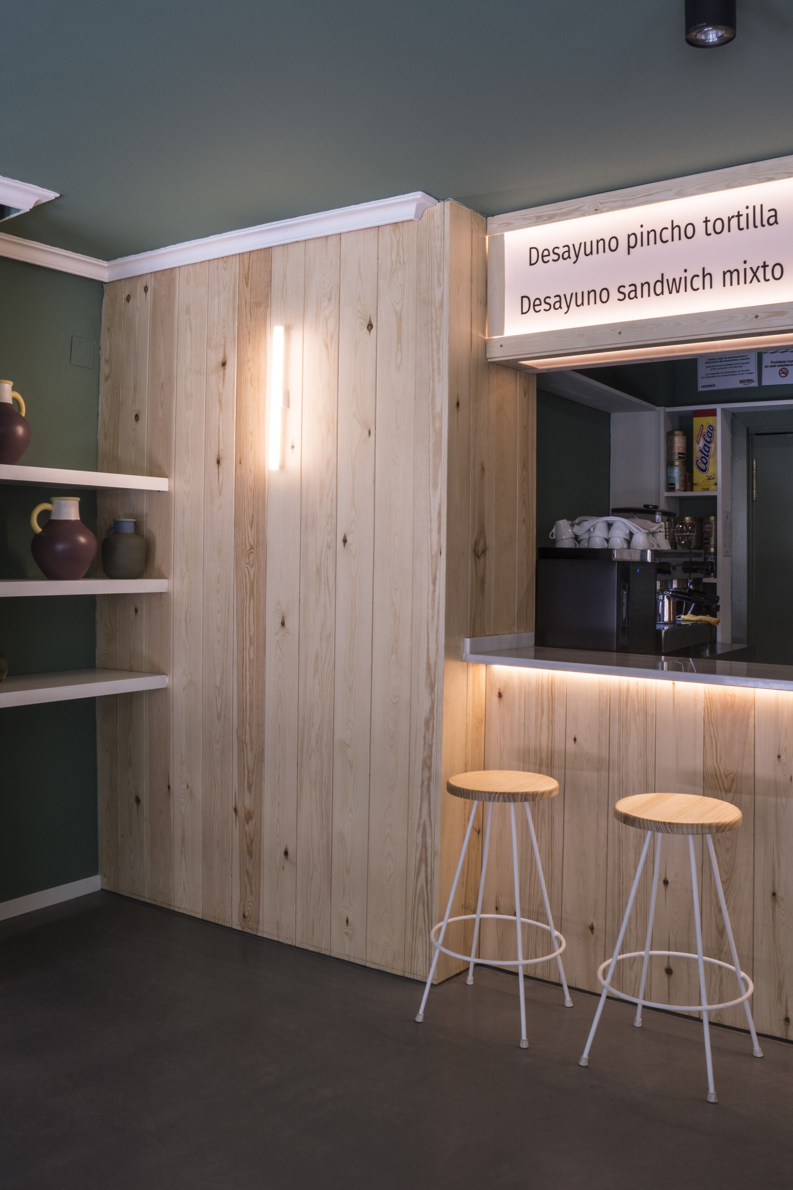 A kitchen with wooden cabinets, a countertop, and two stools. A sign above the counter reads "Desayuno pincho tortilla" and "Desayuno sandwich mixto".
