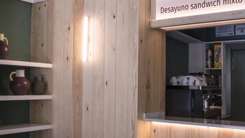 A kitchen with wooden cabinets, a countertop, and two stools. A sign above the counter reads "Desayuno pincho tortilla" and "Desayuno sandwich mixto".