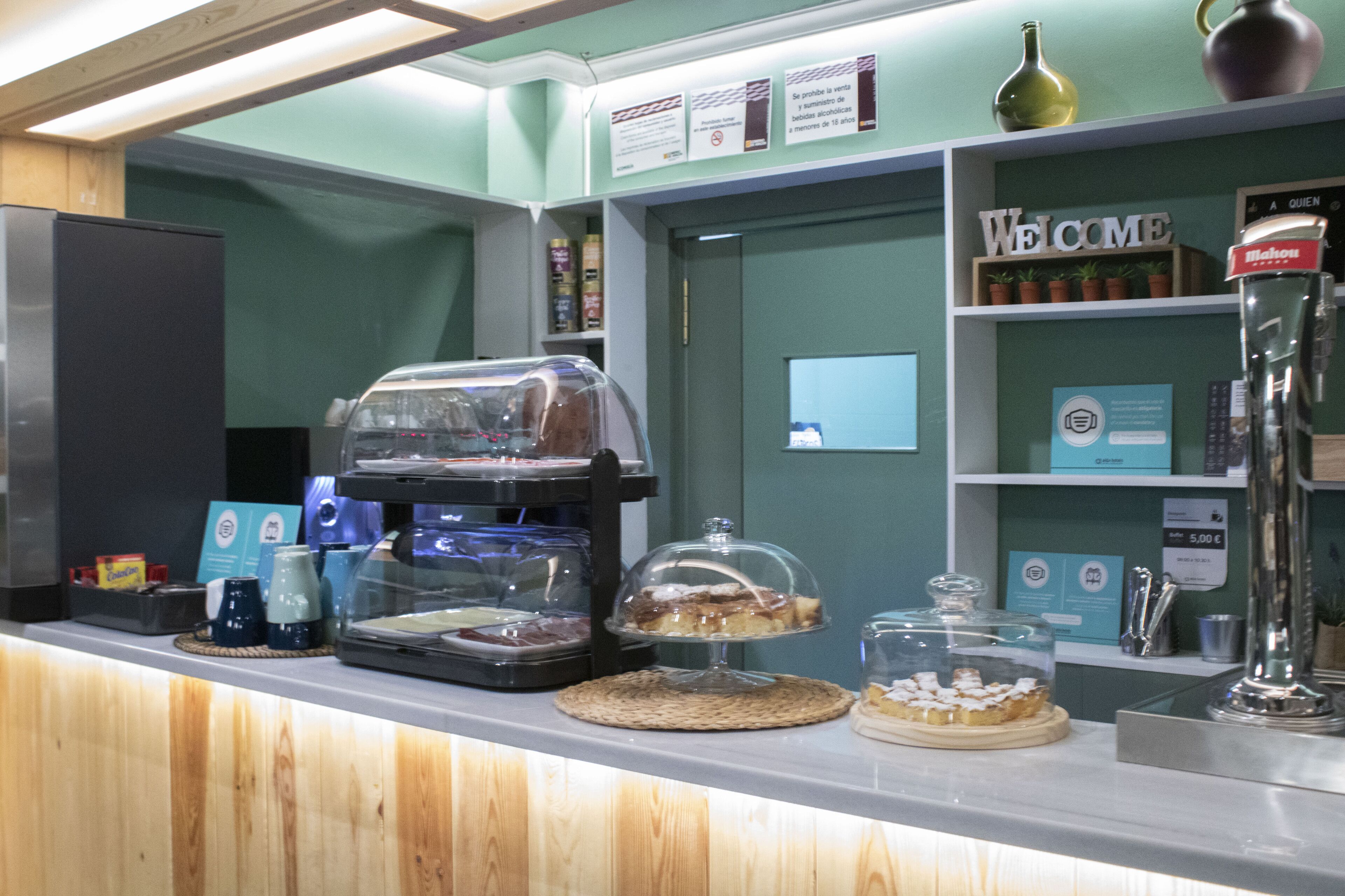 Mostrador de una panadería con diversos productos horneados, una máquina de café y un letrero de bienvenida.
