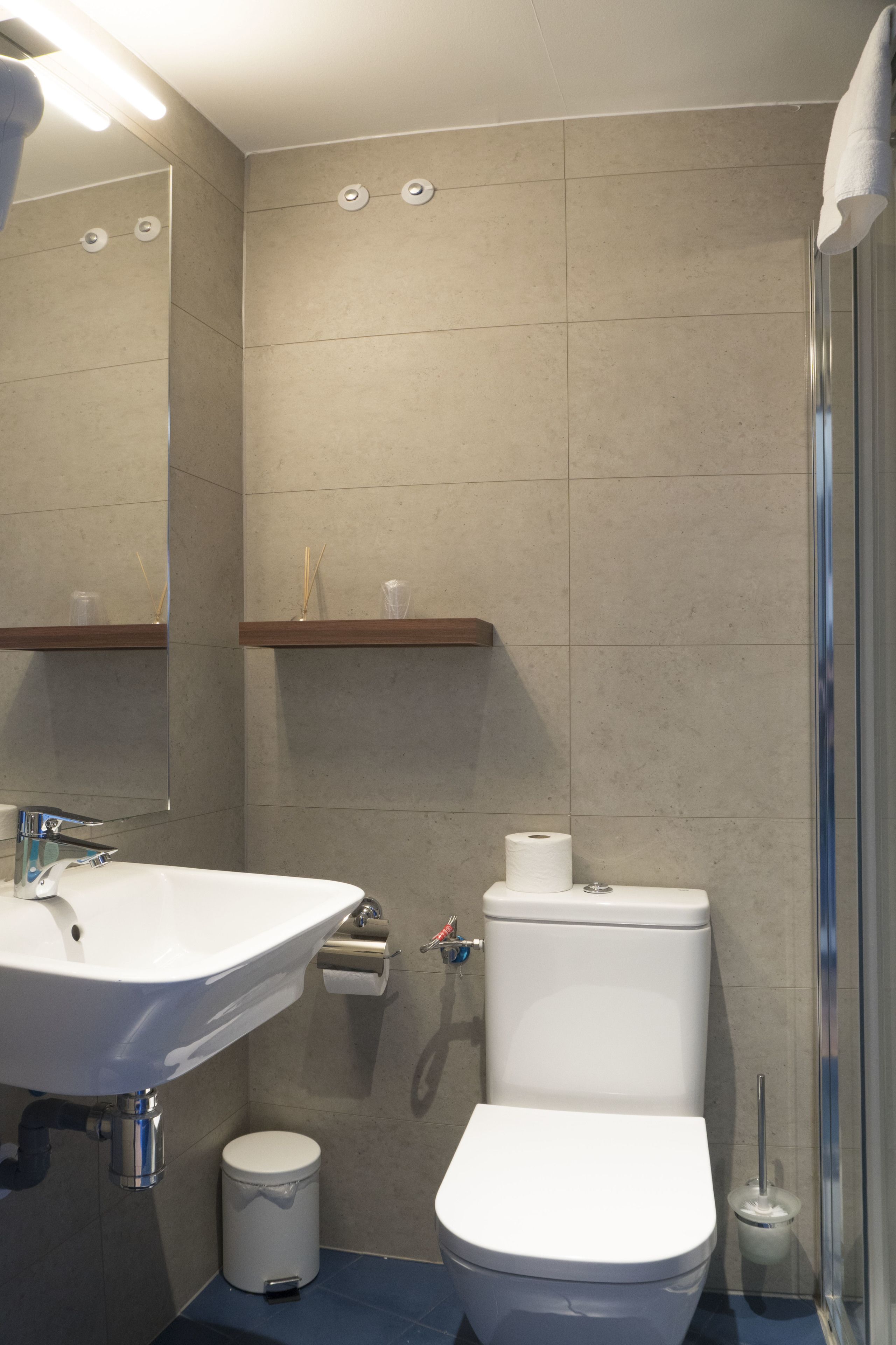 A modern bathroom with a white sink, toilet, and shower. There are shelves above the sink and a trash can next to the toilet.
