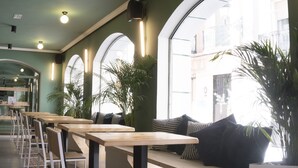 A modern cafe with long wooden tables and chairs, large windows, and potted plants. - Alda Jolio Jaca (Jaca)