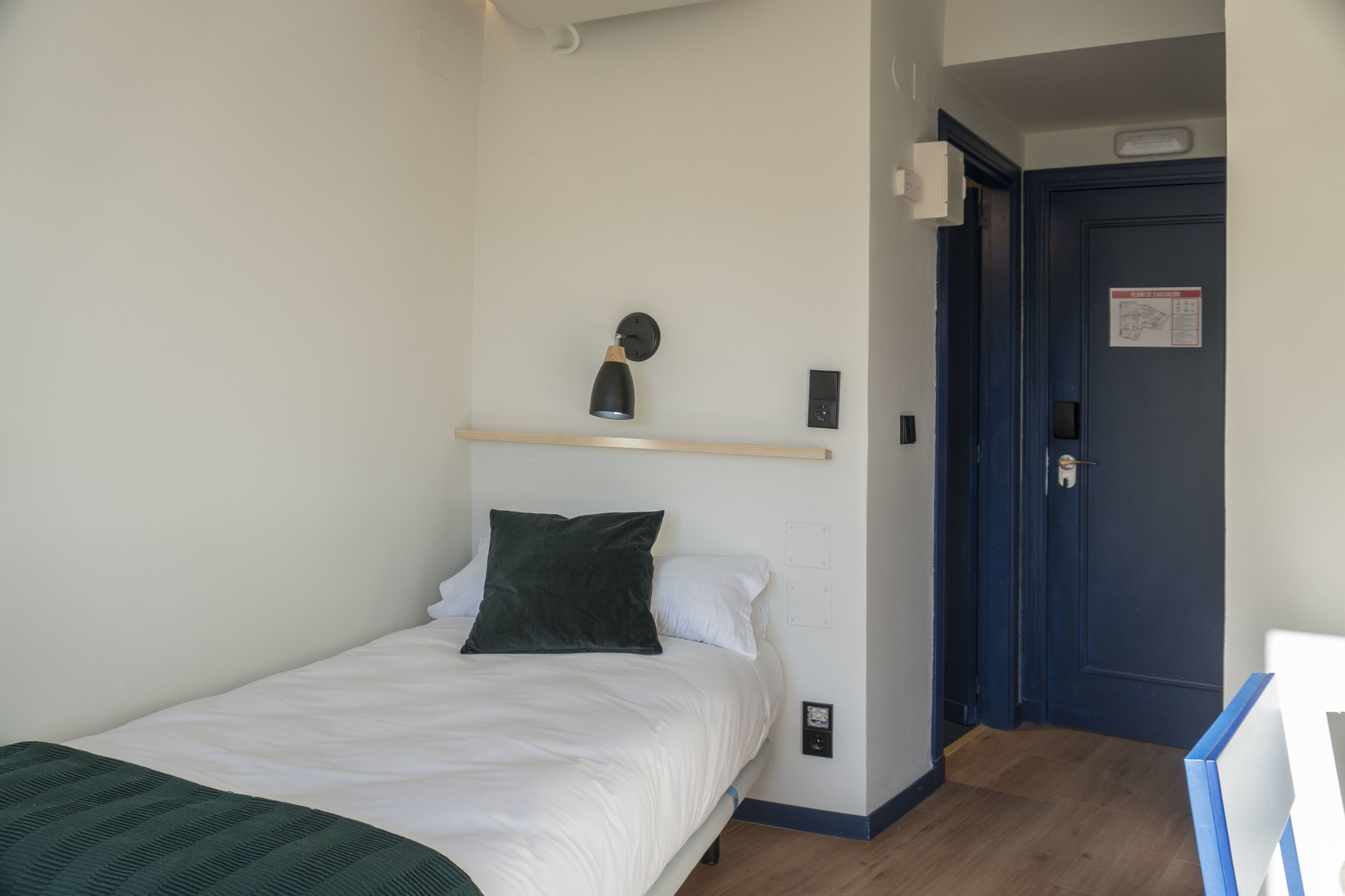 Een slaapkamer met een bed, een bedlamp, een wandplank, een deur met een bordje en een lichtschakelaar.