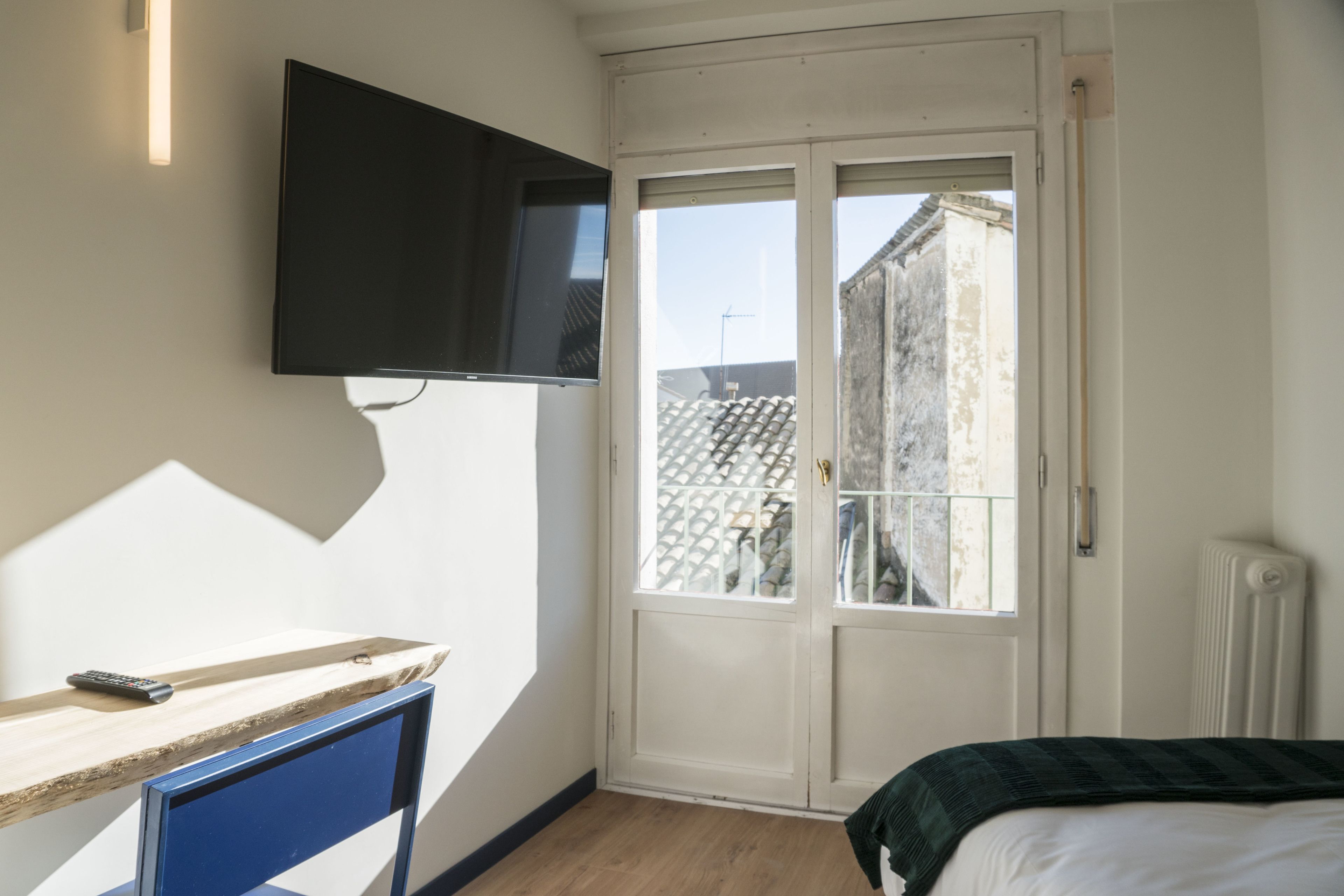 Un dormitorio con cama, televisor montado en la pared, ventana con vista a edificios y un escritorio de madera con control remoto.