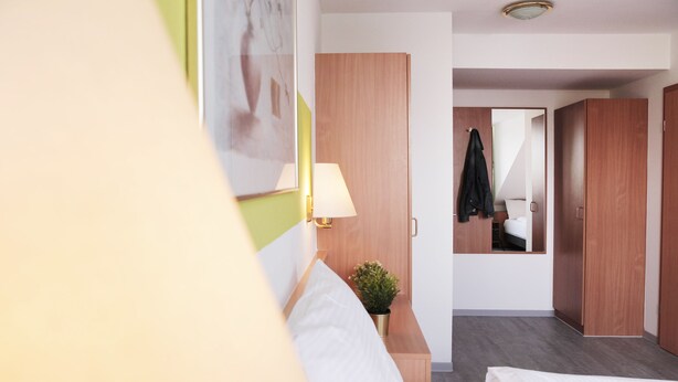 A hotel room with a bed, a bedside lamp, a small plant, and a mirror on the wall. - bp24 Hotel Aachen (Aachen)