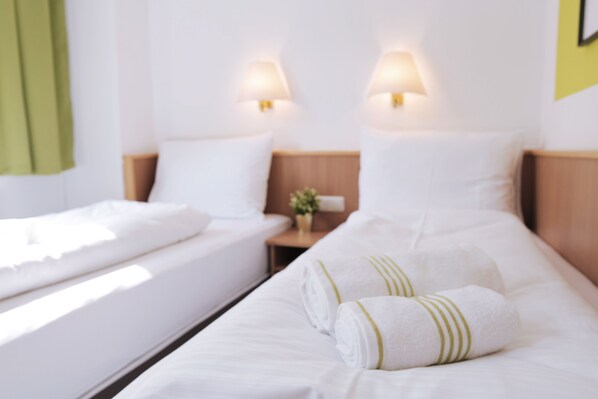 Quarto de hotel com duas camas, cada uma com roupa de cama e almofadas brancas, e duas luminárias fixadas na parede.