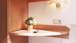Una esquina de una habitación con un estante de madera, una planta en maceta, un control remoto y una lámpara de pared.